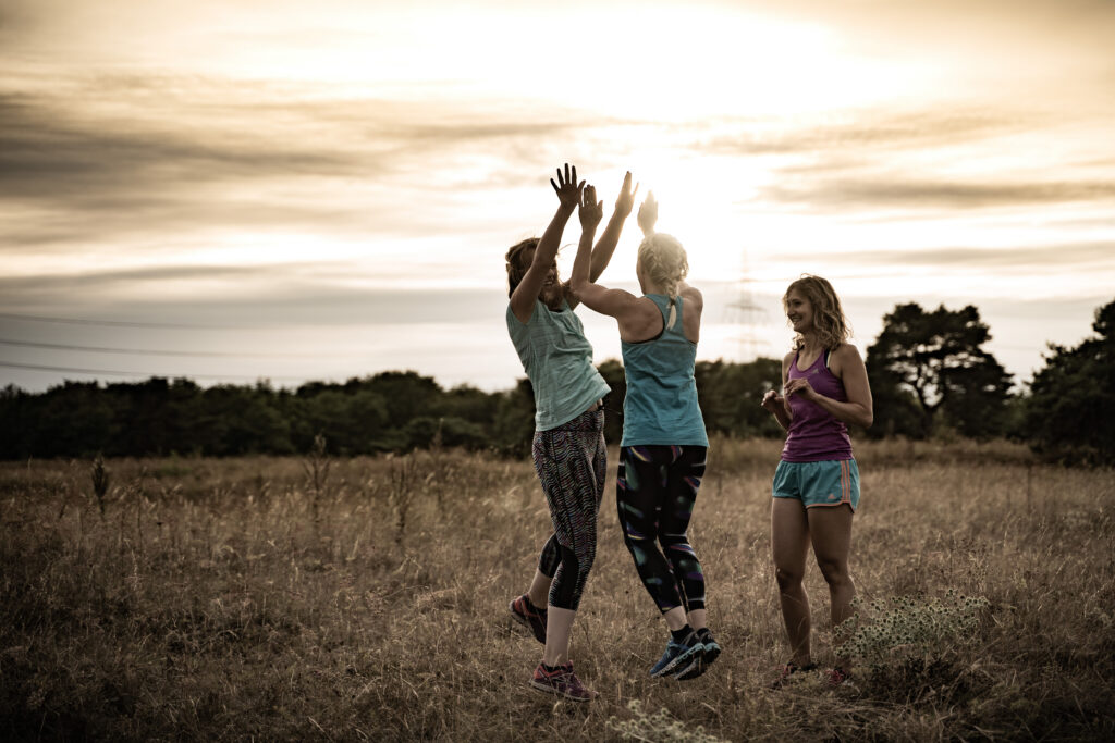 Laufen anfangen – zwei Läuferinnen springen mit High-Five bei Sonnenaufgang, Naturtraining, gemeinsamer Trainingseinstieg
