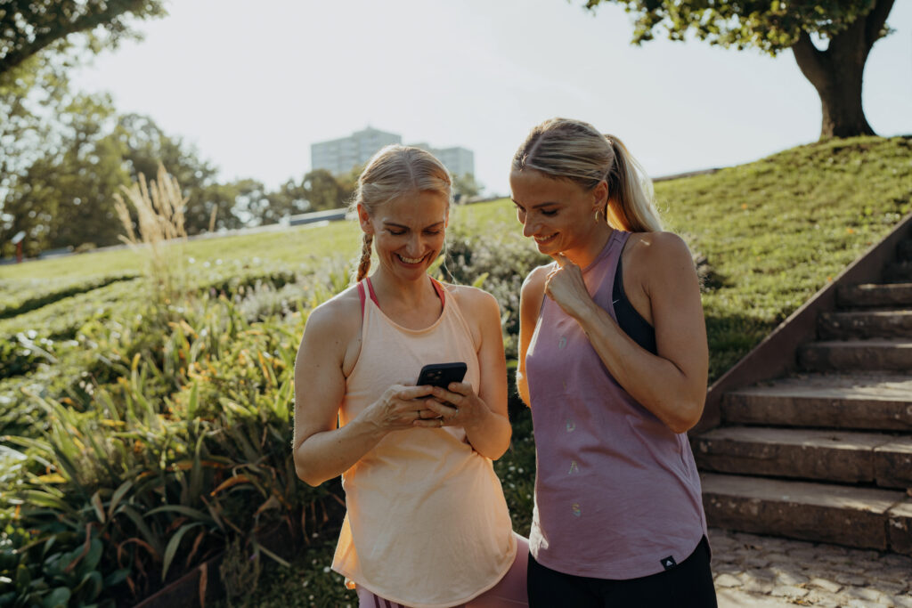 Intervalltraining – zwei Läuferinnen checken Trainingsmetriken auf dem Smartphone, Pause zwischen Intervallen im Park