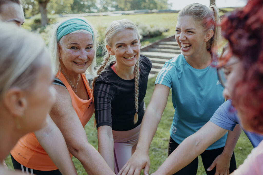 Intervalltraining – Läuferinnengruppe lacht zusammen nach Trainingseinheit, verschiedene Fitness-Level im Team, Park-Setting