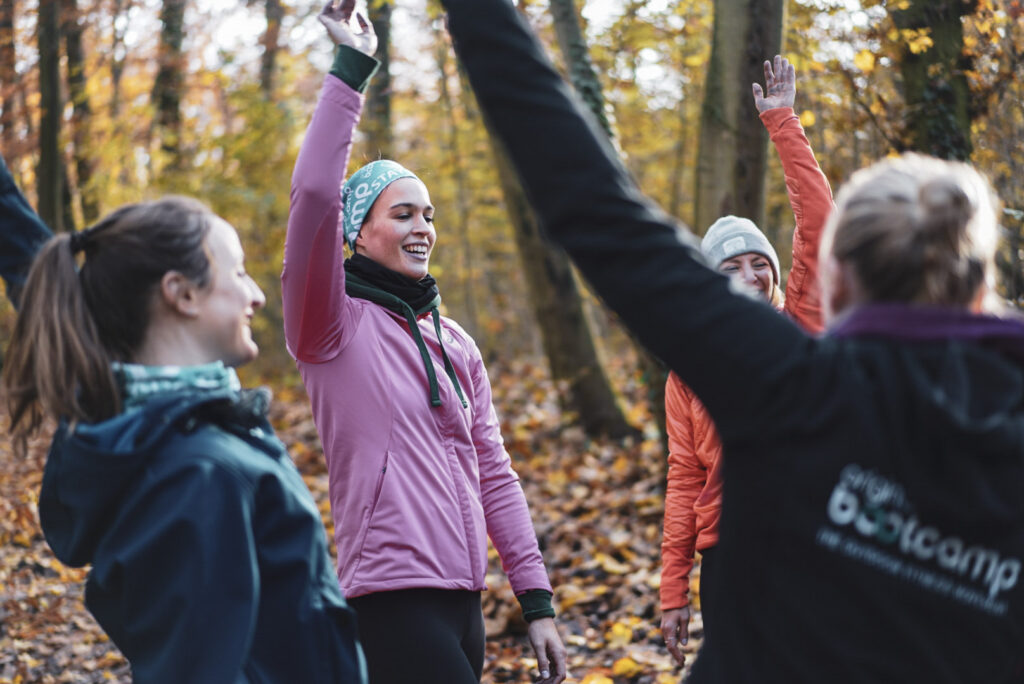 Motivation beim Laufen – Läuferin in lila Jacke jubelt mit erhobenen Armen im Herbstwald, Gruppe feiert zusammen beim Outdoor-Training