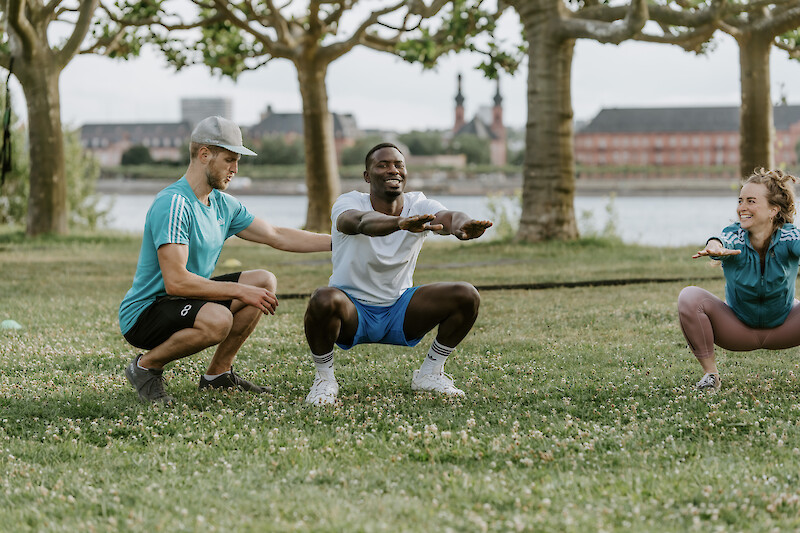 Outdoor Gym Kniebeugen im Park – Trainer begleitet zwei Teilnehmende bei tiefen Squats auf einer Wiese mit Rheinblick.