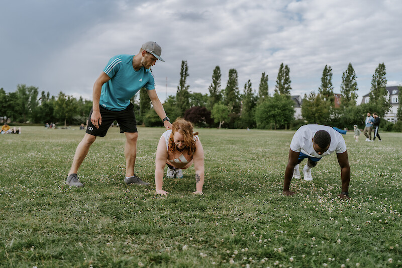 Zirkeltraining Outdoor: Trainer unterstützt zwei Teilnehmende bei einer Plank-Übung.