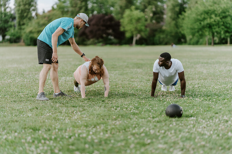 Trainer motiviert Teilnehmende bei Liegestützen auf der Wiese – Zirkeltraining im Freien zur Förderung von Kraft, Stabilität und Muskelaufbau.